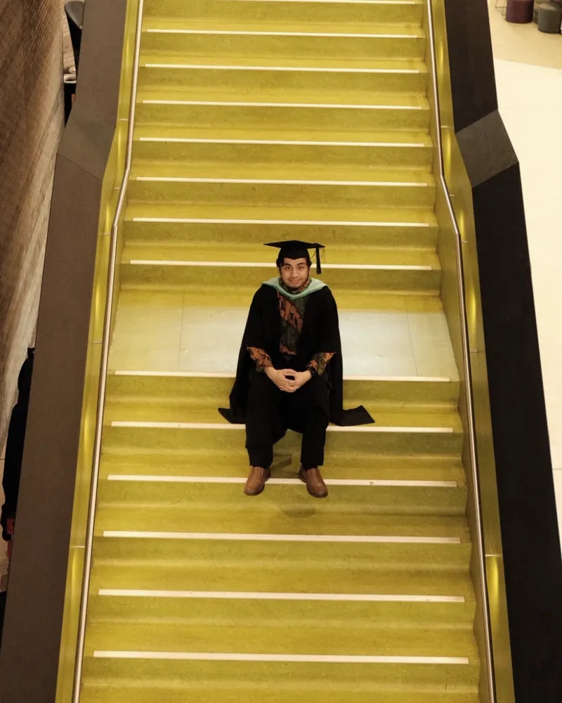Syarif in a graduation gown and cap, sitting on a wide staircase seen from above