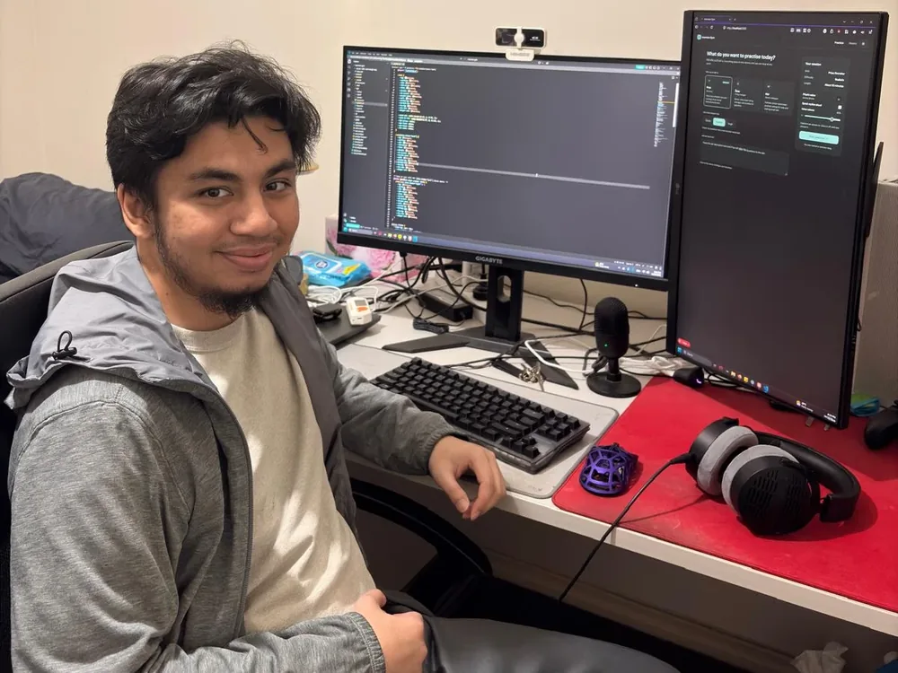 Syarif at his desk, smiling, with a code editor on one monitor and a project open on a vertical second monitor